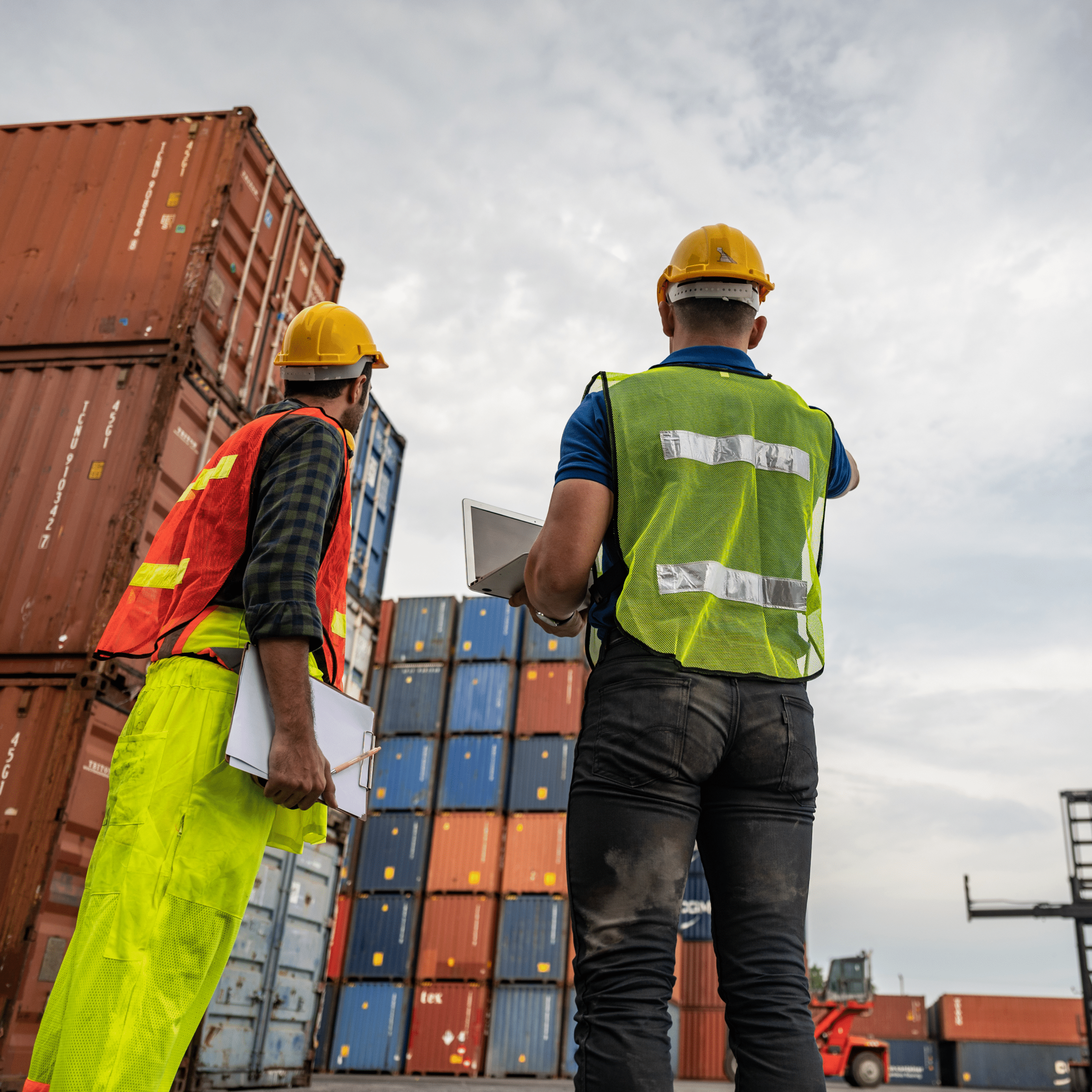 Two terminal workers in safety gear overseeing container stacks at an inland terminal, coordinating operations with a tablet and paperwork.